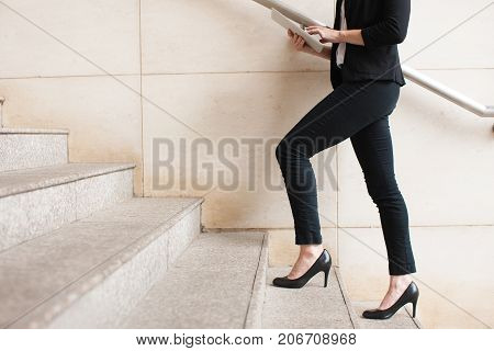 Portrait of young Caucasian businesswoman wearing high-heeled shoes walking upstairs and using digital tablet. Business concept