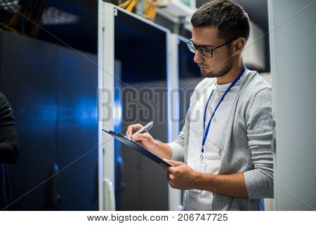 Side view portrait of young network engineer writing data on clipboard while working with servers in data center