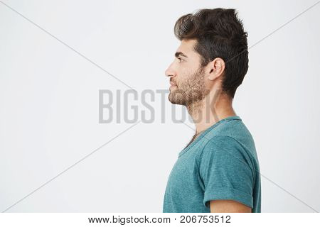 Half-face studio portrait of beautiful spanish man, in casual blue t-shirt, with trendy hair and beard, showing his profile and looking to the left. Copy space