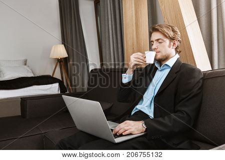 Relaxed handsome businessman with sylish hairstyle and beard sitting in hotel room, drinkig coffee, working on new startup project. Comfortable work place.