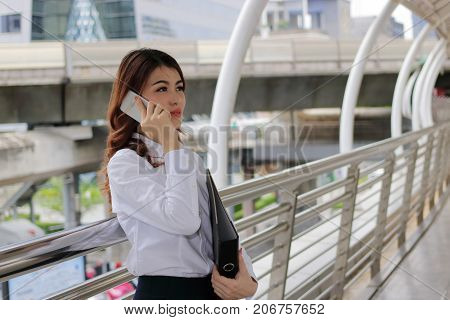 Young Asian businesswoman talking on phone and holding document folder in her hands at urban city background.