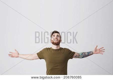 Beautiful bearded european man with tatooed arm and dark hair in casual olive t-shirt spreading hands widely, looking upside, being sutisfied and carefree. Guy feeling free after month of hard work