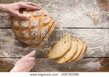Baking background. Hands of baker cutting bread loaf with knife on rustic wooden table sprinkled with flour.