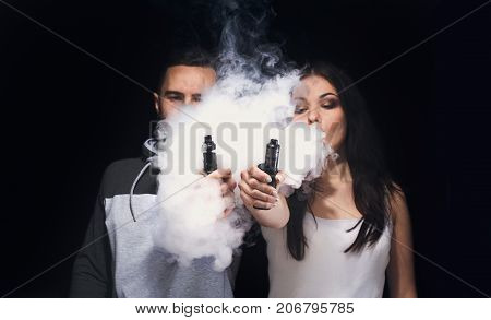 Couple vaping. Unrecognizable young man and woman in the clouds of smoke showing their vapes to camera at black studio background. Relationship and vape addiction concept with copy space