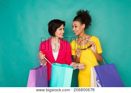 Happy multiethnic girlfriends with shopping bags. Two excited shopaholics boasting with purchases at turquoise sudio background with copy space