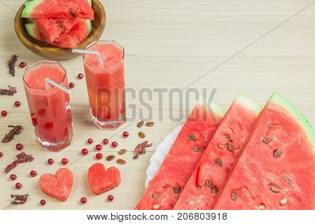 Two hearts, watermelon juice into two glass cups with a straw on a light wooden background, a delicious cocktail, a plate with slices of ripe watermelon on the table. Red berries, cranberries and brown seeds.