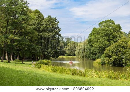 Garden landscape : lake is surrounded by a green lawn and old deciduous trees at sunny summer day. Boat with people in the middle of the lake . Old park Schoenbusch ( Schönbuch ) - english landscape garden Aschaffenburg Germany