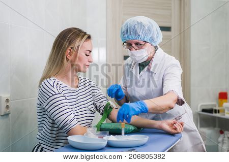 Preparation for blood test with pretty young blond woman by female doctor in white coat medical uniform on the table in white bright room. Nurse pierces the patient's arm vein with needle blank tube