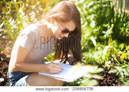Attractive young artist works on plein air drow in pencil. Wearing sunglasses smiling. Enjoying painting on sunner day. Art education art college.