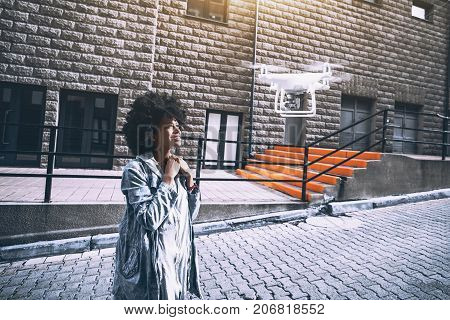 Beautiful cheerful young biracial curly lady posing in front of mobile camera of flying drone in urban settings while making online report for subscribers of her video channel