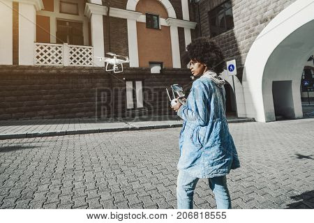 Pensive young biracial curly lady is driving white modern drone using remote controller having screen for showing video from quadrupter's camera while standing on pavement of urban street on sunny day