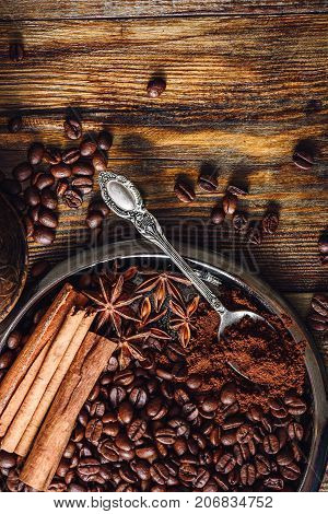 Coffee Beans with Spooonful of Ground Coffee Cinnamon Sticks and Star Anise on Plate. Some Beans Scattered on Wooden Table. View from Above. Vertical Orientation.