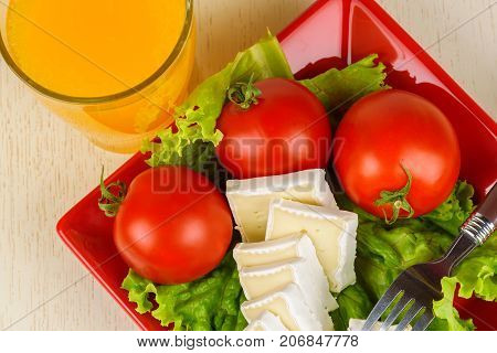 Tomatoes And Cheese Slices Lie On A Sheet Of Fresh Salad In A Red Plate. A Glass Of Orange Juice Sta