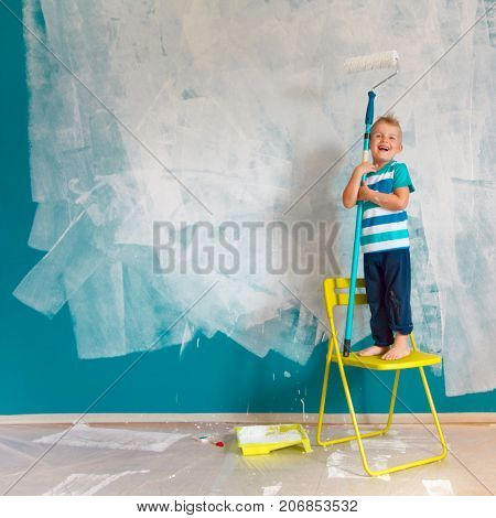 The Child Paints The Blue Wall With A Roller. The Boy Holds A Large Brush For Painting. Home Repairs