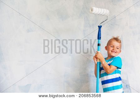 The Child Paints The Blue Wall With A Roller. The Boy Holds A Large Brush For Painting. Home Repairs