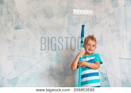 The Child Paints The Blue Wall With A Roller. The Boy Holds A Large Brush For Painting. Home Repairs