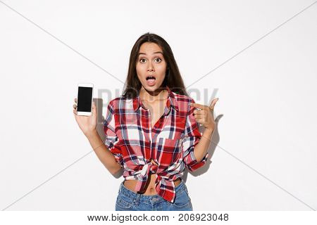 Amazed young pretty girl in checkered shirt showing thumb up gesture and holding smartphone, isolated on white background