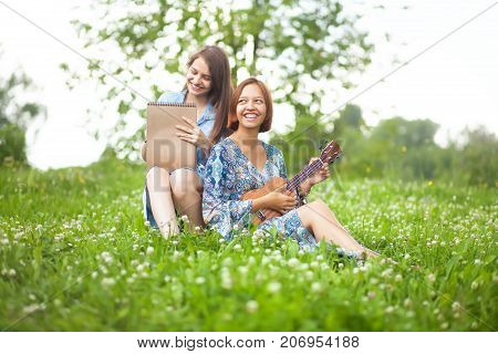 Two talented student girls sisters enjoing their hobby. Drawing on plein air plaing on ukulele. Smiling and laughing having fun together. Sister friendship and relationship