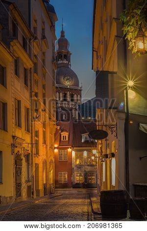 Riga, Latvia. View Of Narrow Cobbled Kramu Street Of Old Town In Bright Evening Illumination With Ancient Architecture Leading To Dome Cathedral Under Summer Blue Sky.