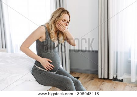 Attractive young pregnant blonde woman feeling sick while sitting on bed at home