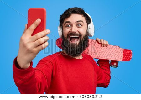 Cheerful Bearded Hipster In Trendy Red Outfit And Modern Headphones Holding Skateboard On Shoulder A