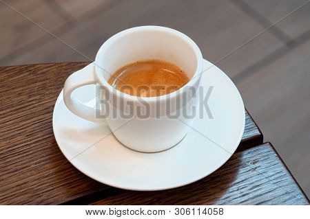 Italian Espresso In A White Ceramic Cup With Saucer On A Brown Wood Table. Brurred Background.