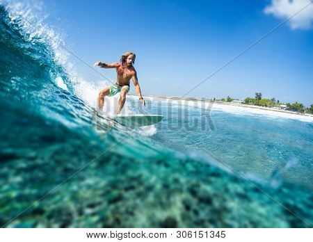 HIMMAFUSHI / MALDIVES - MARCH 08, 2019: Caucasian long haired surfer rides the tropical ocean wave with green island on the horizon and some underwater view of the coral reef.Tilt shift effect applied