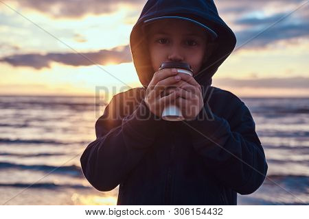 Cute Little Kid Is Enjoying Hot Beverege In Cold Evening At Seaside. There Are Beautiful Sunset Behi