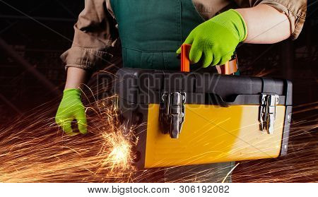 Photo of a worker in green overall outfit with protective gloves holding yeallow toolbox with sparks torso view on black construction area background.
