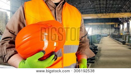 Photo of a worker in orange jacket outfit with gloves holding protective orangle helmet torso view on factory area background.