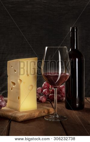 A Glass Of Red Wine With A Bottle, A Large Piece Of Cheesse, And Grapes On A Dark Rustic Background,