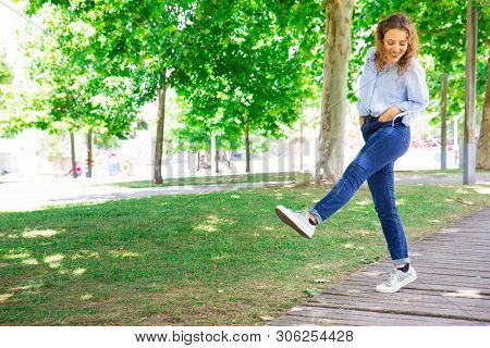 Positive Woman Ambling In Park. Cheerful Curly-haired Girl In Jeans Walking Straight Leg Raise Outdo
