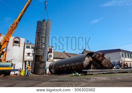Crane Is Bringing Down Heavy Metal Silo In Industrial Complex. Worker Is Helping To Manage Huge Cons