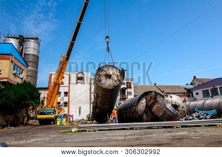 Crane Is Bringing Down Heavy Metal Silo In Industrial Complex. Worker Is Helping To Manage Huge Cons