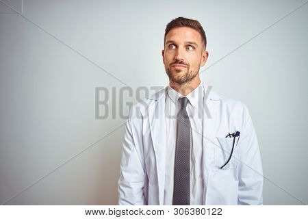 Young handsome doctor man wearing white profressional coat over isolated background smiling looking to the side and staring away thinking.