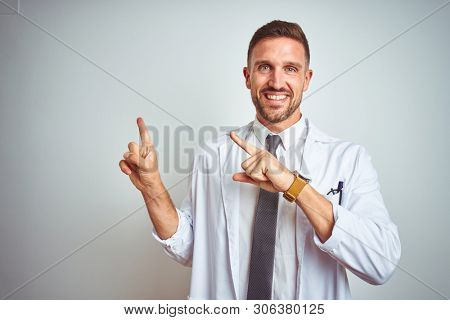 Young handsome doctor man wearing white profressional coat over isolated background smiling and looking at the camera pointing with two hands and fingers to the side.