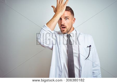 Young handsome doctor man wearing white profressional coat over isolated background surprised with hand on head for mistake, remember error. Forgot, bad memory concept.