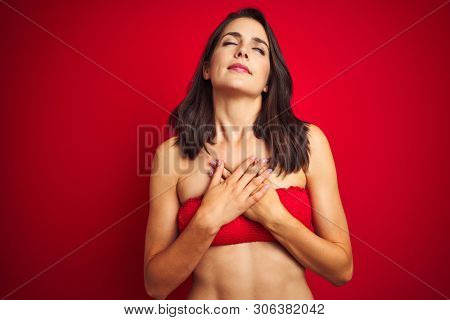 Beautiful woman wearing bikini swimwear over red isolated background smiling with hands on chest with closed eyes and grateful gesture on face. Health concept.