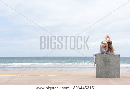Woman Yogi Doing The Pigeon Pose On A Stone Cube On The Beach With The Sea In The Background
