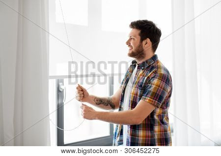 people concept - young man opening or closing roller blind on window at home