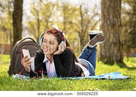 Young Beautiful Girl Lying On A Blanket On The Grass In The Park, Listening To Music And Looking At 