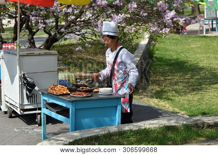 Pyongyang, North Korea - May 1, 2019: Female Seller Of Roast Meat Shown In The Taesongsan Funfair. I