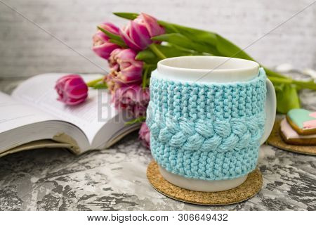 White Cup Of Tea In A Knitted Blue Case With An Open Book And With Pink Flowers Tulips And Sweets, W