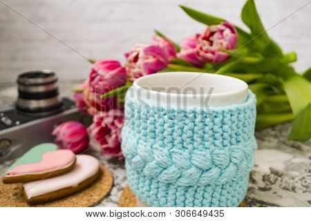 A White Cup Of Tea In A Knitted Blue Case With An Old Camera And Pink Flowers Tulips And Sweets, Gin
