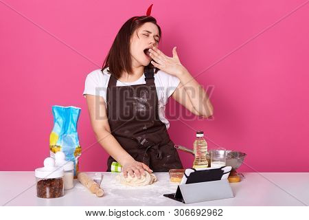 Horozontal Shot Of Sleepy Housewife Dressed Kitchen Apron Dirty With Flour, Whitet Shirt, Red Headba