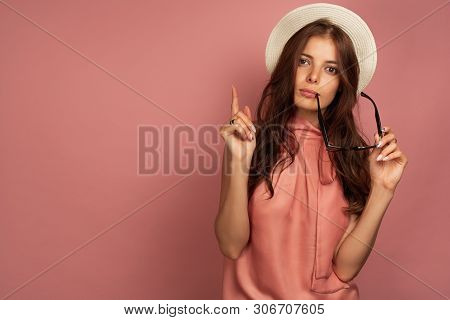 Young Dark-haired Woman With Straw Hat Points Up Holding Her Sunglasses, Pink Background