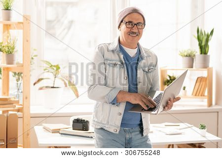 Mature smiling man in denim jacket, blue jeans and pullover standing by desk and typing on laptop keypad in office