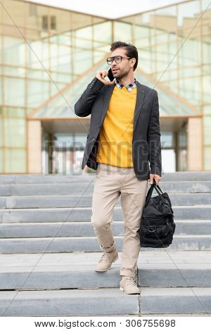 Young business traveler with luggage bag moving downstairs while talking by smartphone in urban environment