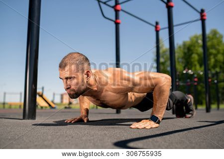 Young muscular shirtless athlete keeping body over sportsground while doing push-ups during outdoor workout