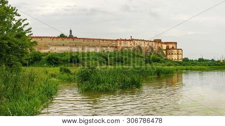 Ancient Brick Medzhybizh Fortress Of The 14-16th Centuries With River On A Foreground In Cloudy Weat